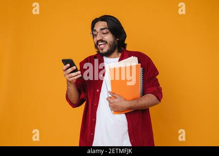 Excited young indian man student holding textbooks isolated over yellow background, using mobile phone Stock Photo
