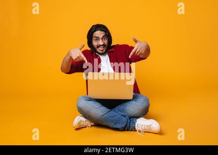 Happy indian man using laptop computer isolated over yellow background, sitting on a floor, pointing at laptop Stock Photo