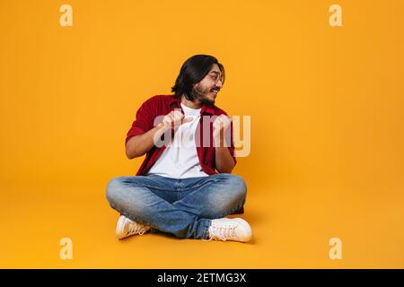 Attractive young indian man presenting copy space isolated on yellow background, sitting with legs crossed Stock Photo