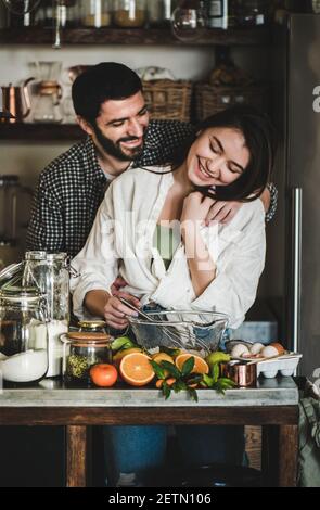 Young loving couple cooking together in kitchen. Young caucasian man and brunette woman hugging, flirting, laughing and making dough for baking citrus cake. Valentines Day, home hobbies concept Stock Photo
