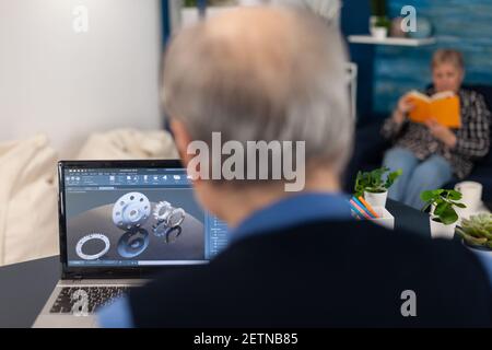 Engineer designed gears parts using professional software. Experienced engineer working from home office on mechanical project using laptop computer technology and wife is reading a book on sofa. Stock Photo
