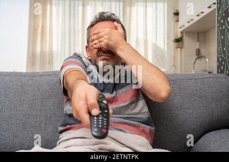 Adult man making scared afraid gesture covering eyes using hands watching horror movie on tv changing channel with remote control as at home pandemic Stock Photo