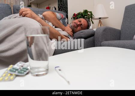 Ill adult man touching neck as sore throat cold flu symptom resting on sofa at home keeping medicine pills water and thermometer on table Stock Photo