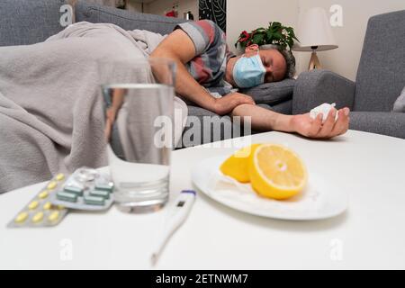 Sick male model wearing surgical or medical protective mask to prevent contamination sleeping on sofa cold sars covid19 treatment pills water thermome Stock Photo