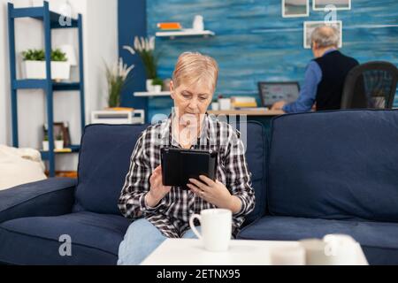 Middle aged lady relaxing on sofa reading on tablet pc anjoying content. Elderly woman using moder technoloy tablet pc in home living room and husband working on laptop computer Stock Photo