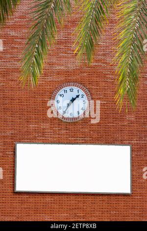 Clock on the red brick facade of a building with palm fronds on the top side and a white sign with copy space. Concepts passing time or being in a hur Stock Photo