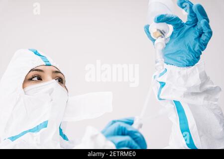 Low angle of female doctor in protective mask and uniform standing in white medical room and using dropper during COVID 19 epidemic Stock Photo
