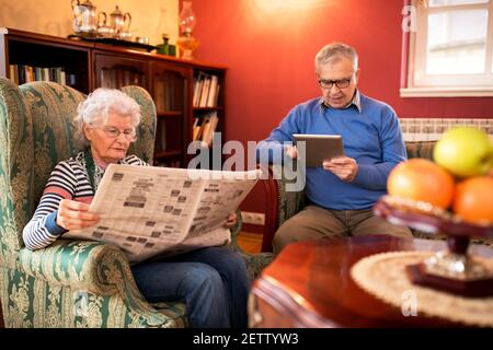 Senior couple relax at home while reading newspaper and using tablet Stock Photo