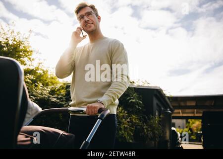 Father pushing at baby stroller talking on mobile phone outdoors. Man on paternity leave taking his baby out in pram and talking on mobile phone. Stock Photo