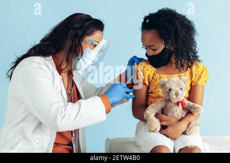 Female doctor wearing protective face mask, gloves and shield injecting vaccine into arm of girl in mask during pandemic. Small girl holding her teddy Stock Photo