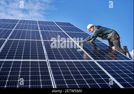 Horizontal snapshot of beautiful and shiny solar battery surface and male worker wearing a uniform, standing on ladder, installing solar modules on sunny day, low angle view Stock Photo