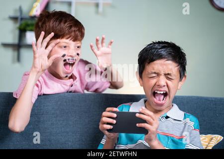 young kid with face painting fools or making prank to his brother while he is busy on mobile by suddenly screaming and shouting from back side at home Stock Photo