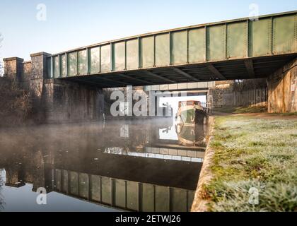 Two old green steel train railway bridges crossing above British canal river reflect in calm still water with old boat passing underneath and towpath Stock Photo
