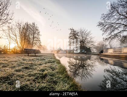 Stunning old canal house boats landscape sunrise in countryside with river and single lone wooden bench. Frost on grass reflection of trees and vessel Stock Photo