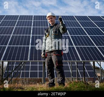 Portrait of workman standing in solar field in front of solar station, showing thumbs up, wearing protective uniform, gloves and helmet, full length. Alternative energy concept. Ecology concept Stock Photo