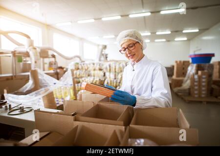 Close up view of a young female pleased worker in sterile cloths packing finished food products in a boxes in food factory storage. Stock Photo