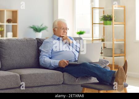 Senior man sitting in the room on the couch with a laptop in his hands and thinking. Stock Photo