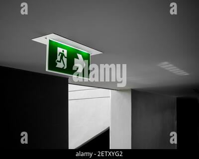 Fire exit sign. Green emergency exit sign hanging from ceiling glowing at the dark corridor near the fire escape door in the building. Stock Photo