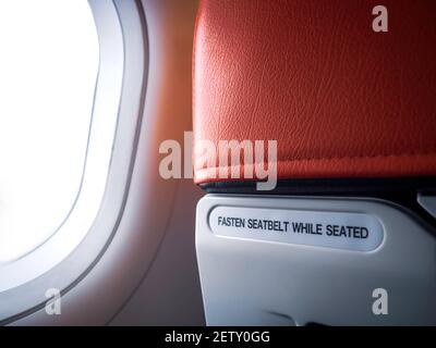 Text message 'FASTEN SEAT BELT WHILE SEATED' behind the red airplane seat near the window in cabin of low cost commercial airline for information to p Stock Photo