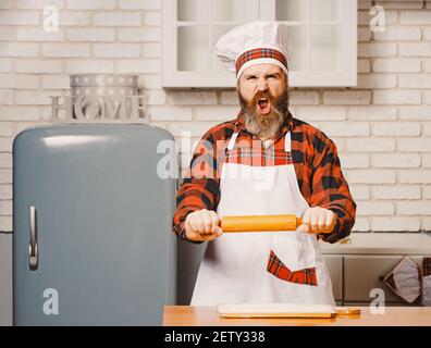 Angry chef bakers man raising rolling pin threateningly in white kitchen. Stock Photo