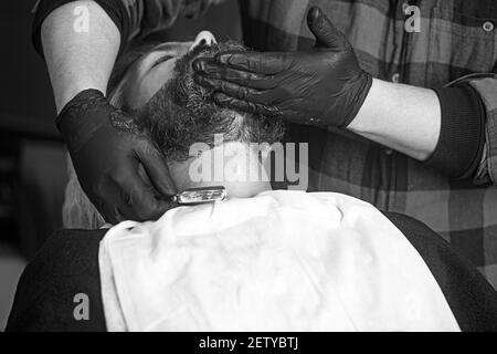 Hand of barber keeping straight razor and cutting trendy style on beard of client. Concept of shaving. Stock Photo
