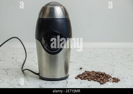 An electric coffee grinder sits on a white table, with roasted coffee beans lying next to it. The concept of freshly brewed coffee Stock Photo