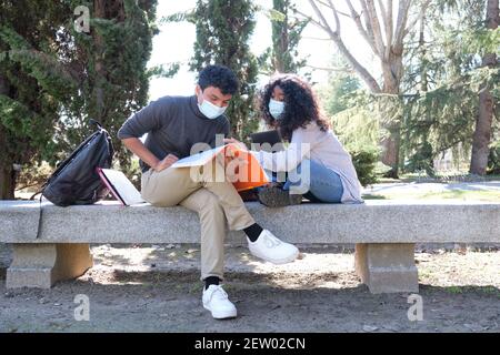 Two latin students wearing protective face mask studying together sitting on a bench outdoor. New normal at university campus. Stock Photo