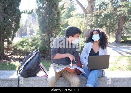 Two latin students wearing protective face mask studying together sitting on a bench outdoor. New normal at university campus. Stock Photo