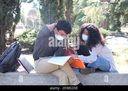 Two latin students wearing protective face mask studying together sitting on a bench outdoor. New normal at university campus. Stock Photo
