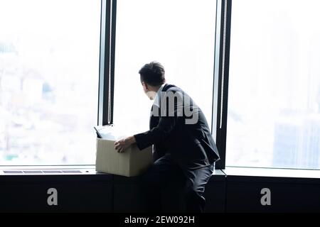 Fired businessman sitting with box of belongings by window Stock Photo