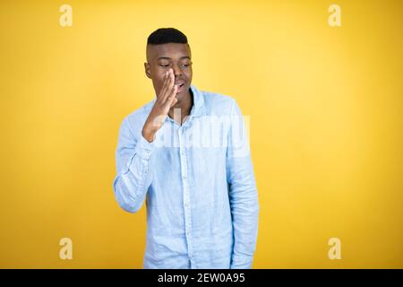 Young african american man wearing a casual shirt standing over yellow background hand on mouth telling secret rumor, whispering malicious talk conver