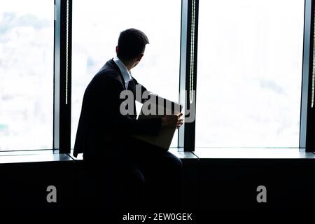 Fired businessman sitting with box of belongings by window Stock Photo