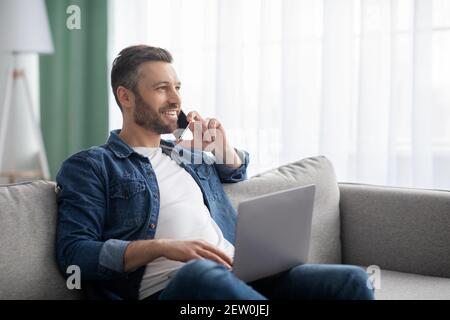Cheerful man enjoying weekend at home, using laptop and phone Stock Photo