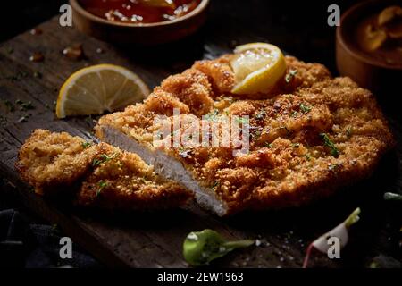 Tasty fried breaded meat under lemon and herbs with sauces on wooden board in restaurant Stock Photo