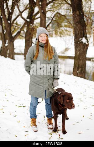 Smiling young woman with brown dog labrador retriever in winter warm outfit walking outdoors in snowy park, full height looking at camera Stock Photo