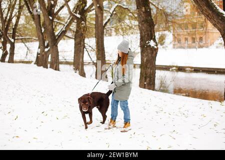 Smiling young woman with brown dog labrador in winter warm outfit walking outdoors in snowy park, full height looking at camera Stock Photo