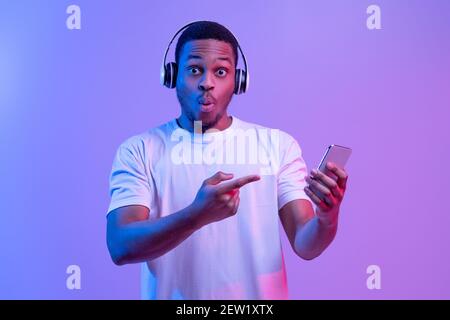 Cool App. Surprised Black Guy In Wireless Headphones Pointing At Smartphone Stock Photo
