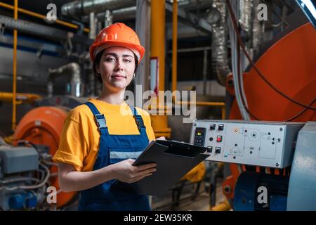 Portrait of female smiling worker in uniform and helmet with a folder and a pen in her hand. In the background-boiler room. Stock Photo