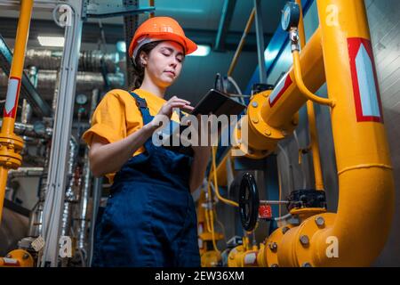 A young female engineer in a uniform and a protective helmet, holding a digital tablet in her hands and conducting an inspection of the equipment. Bot Stock Photo