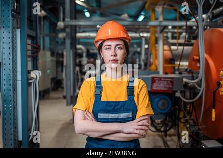 Portrait of a young tired female worker in a work uniform and helmet, arms crossed. In the background is a boiler room. The concept of industrial prod Stock Photo