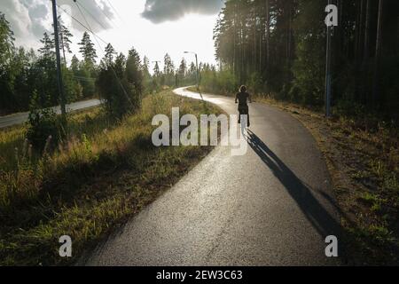 A woman rides a bicycle on a bicycle path near a pine forest and in the rays of the setting sun. Healthy lifestyle concept. Stock Photo