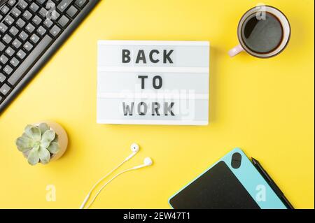 Back to work - text on display lightbox on yellow background workplace. Black keyboard, white earphones, coffee, tablet. Stock Photo