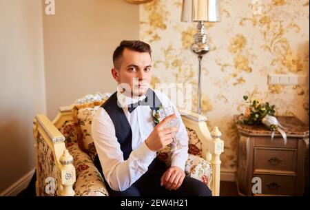 Morning of groom. Beautiful man, groom posing and preparing for wedding. Stylish groom sits on the black chair in studio and holds a glass of whiskey. Stock Photo