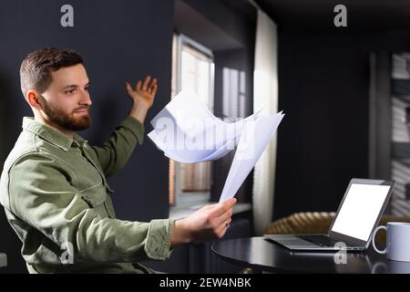 A tired disgruntled caucasian man scatters documents at home, working remotely. Side white space. Stock Photo