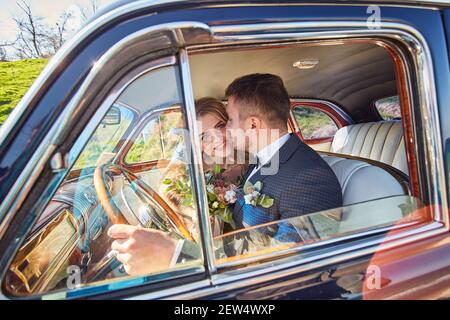 elegant gorgeous bride smiling and handsome stylish groom kissing her in car. unusual luxury wedding couple in retro style. romantic moment Stock Photo