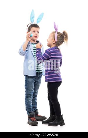 Candid cute little kids siblings wearing bunny ears playing with Easter painted eggs. Full body isolated on white background. Stock Photo