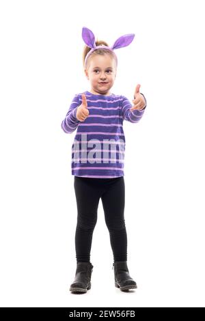 Cute little girl with Easter bunny ears showing thumbs up looking away curious. Full body isolated on white background. Stock Photo