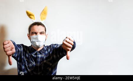 Charming Caucasian man with beard yellow rabbit eared and protective medical mask puts on dislike with both hands. Celebrate Easter in coronavirus yea Stock Photo