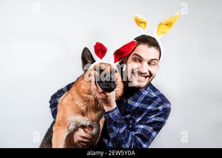 Caucasian man with yellow rabbit ears hugs German shepherd with red plush ears. Creative banner for celebration of Easter. Man and dog on white backgr Stock Photo