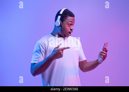 Amazed Black Guy In Wireless Headphones Pointing At Smartphone In His Hands Stock Photo